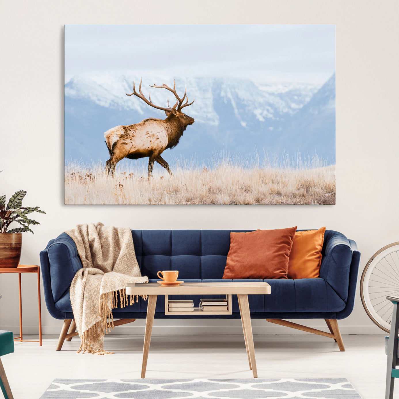 An elk walking on snowy grassland with majestic mountains in the background, ideal for rustic lodge or cabin wall decor.