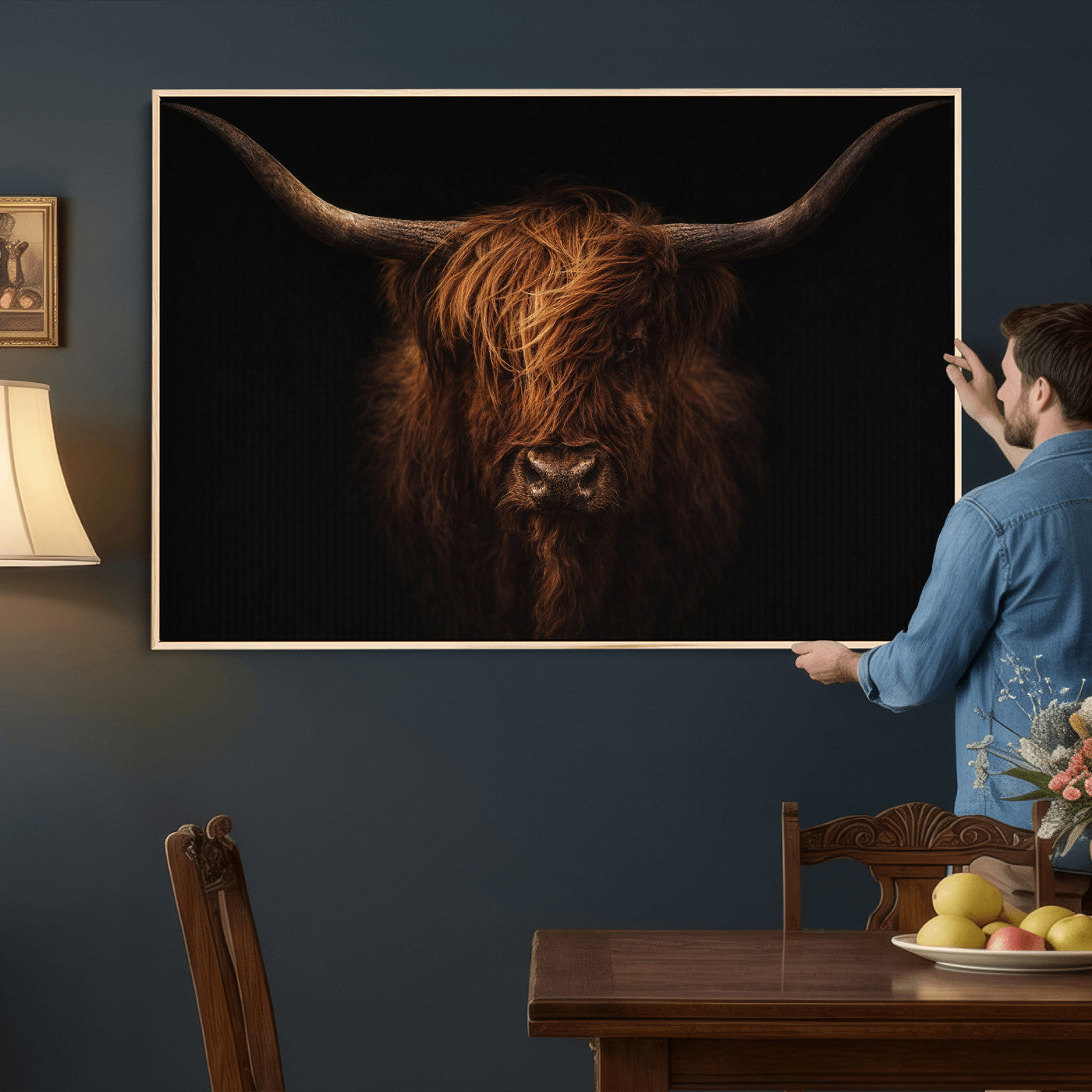 Man hanging a framed picture of a highland cow on a wall in a room with a table and chair.