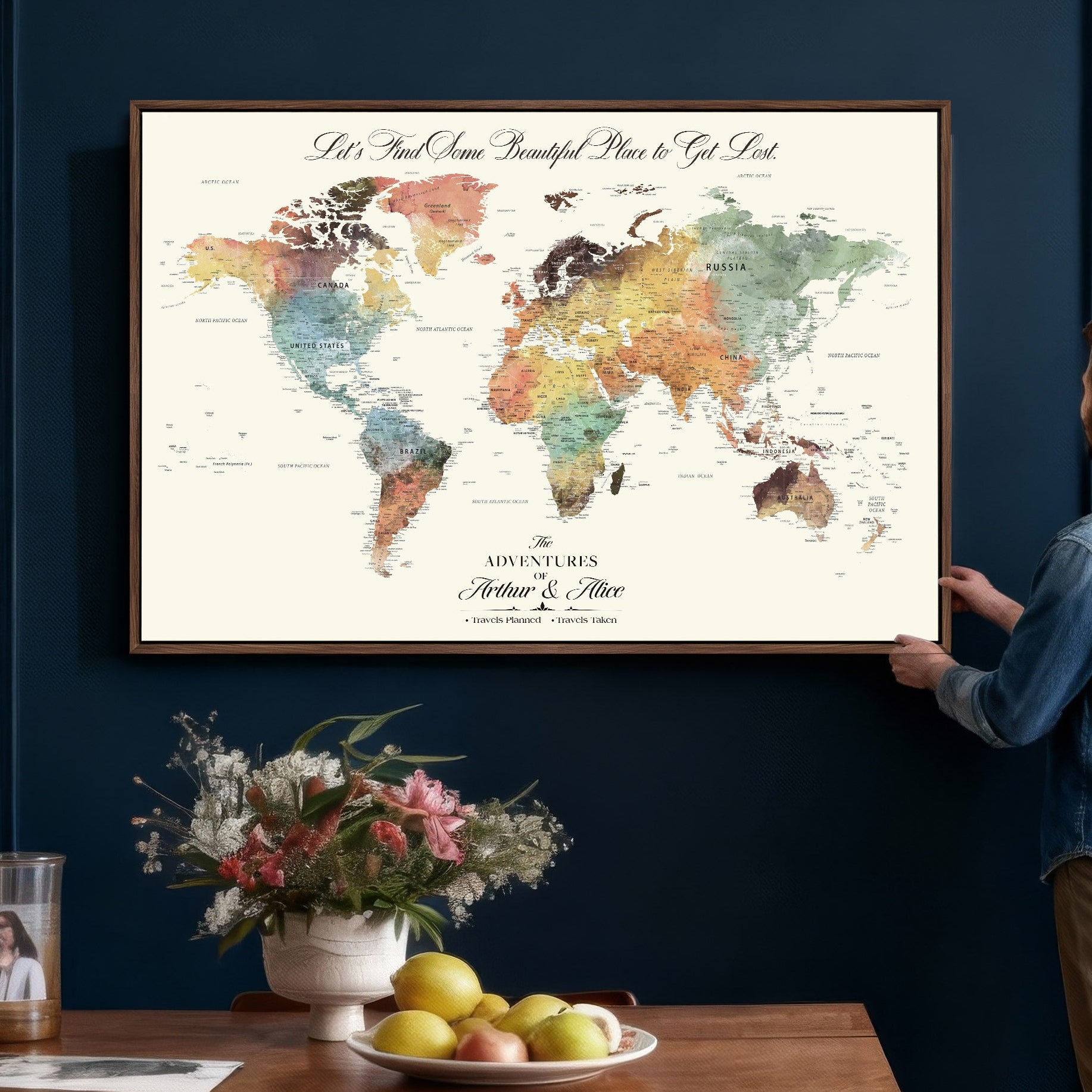 Person hanging a framed world map on a blue wall with a table and flowers in the foreground.