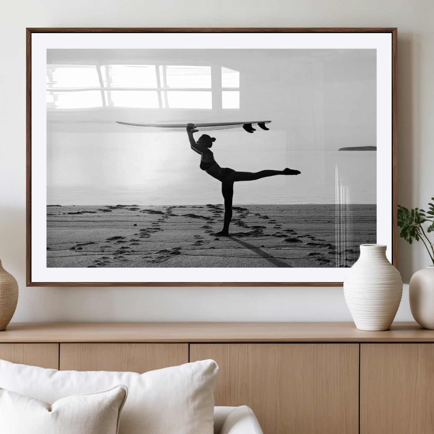 A woman balances a surfboard on her head while striking a yoga pose on the beach, silhouetted against a calm ocean horizon.