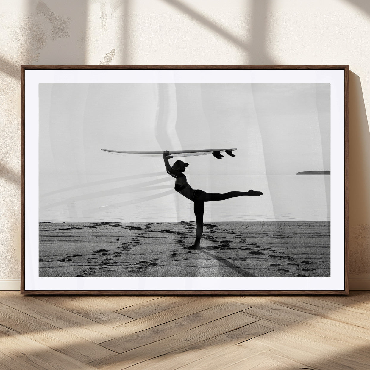 A woman balances a surfboard on her head while striking a yoga pose on the beach, silhouetted against a calm ocean horizon.