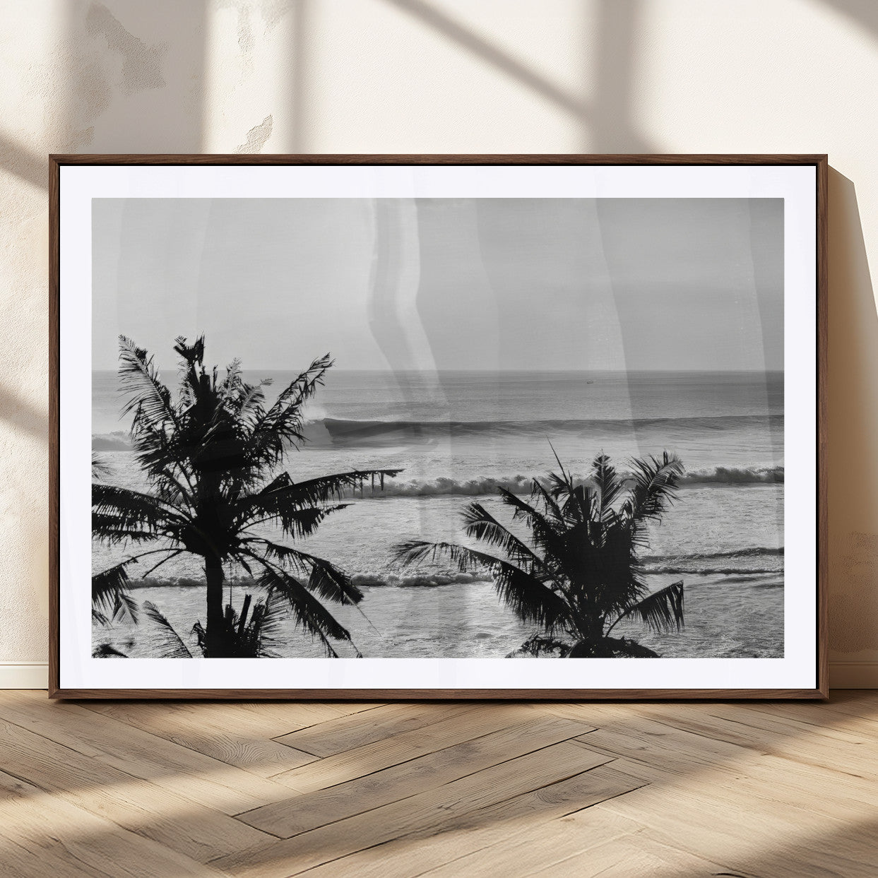 Black and white view of ocean waves crashing on a tropical coastline, framed by silhouetted palm trees under a vast, open sky.