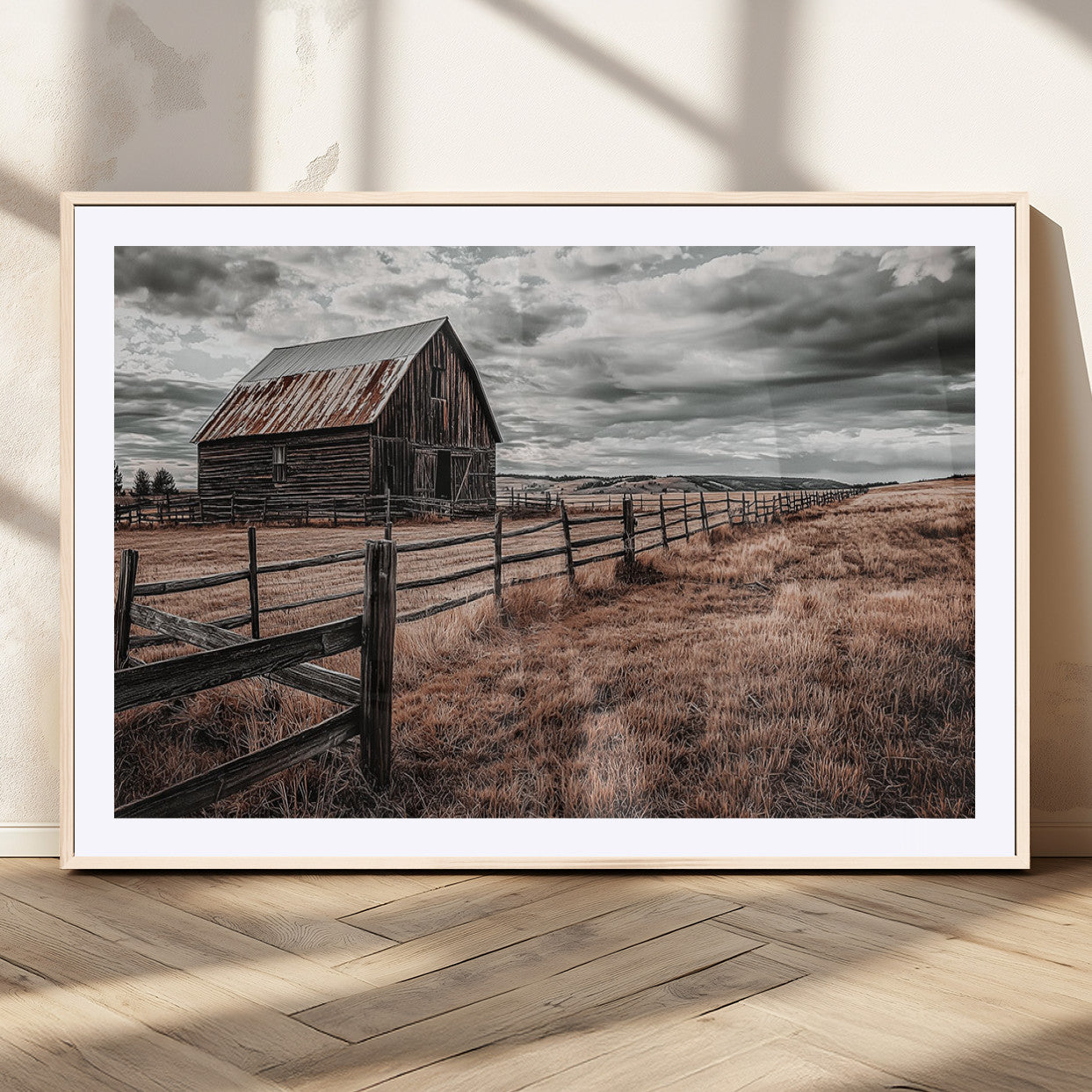 "Black-and-white photograph of a rustic barn with a weathered roof, surrounded by a wooden fence and vast golden fields under a moody sky."