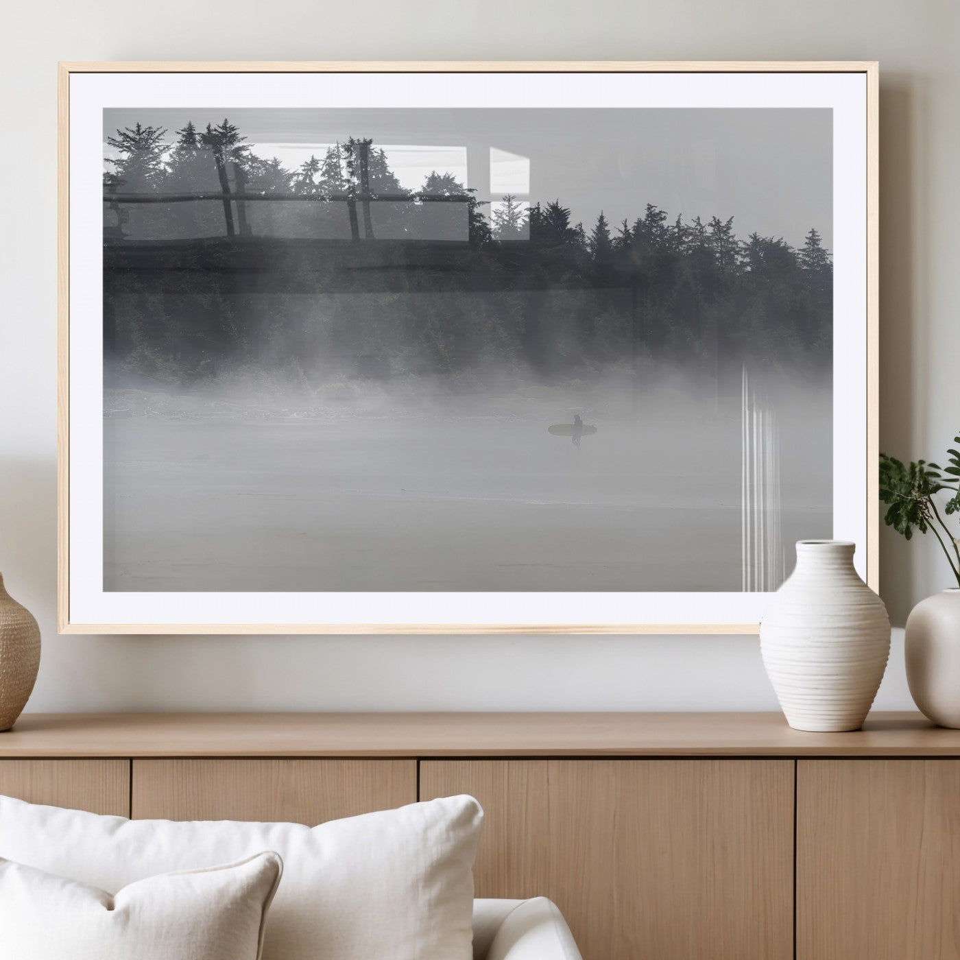 A lone surfer carrying a board through a misty shoreline, surrounded by dense trees and fog, creating a peaceful and mysterious coastal scene.