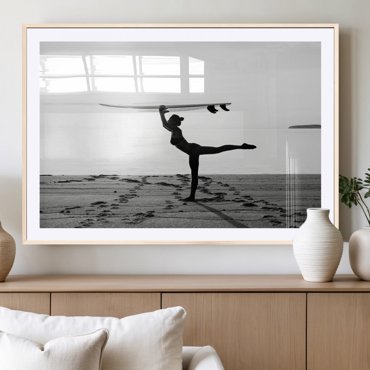 A woman balances a surfboard on her head while striking a yoga pose on the beach, silhouetted against a calm ocean horizon.