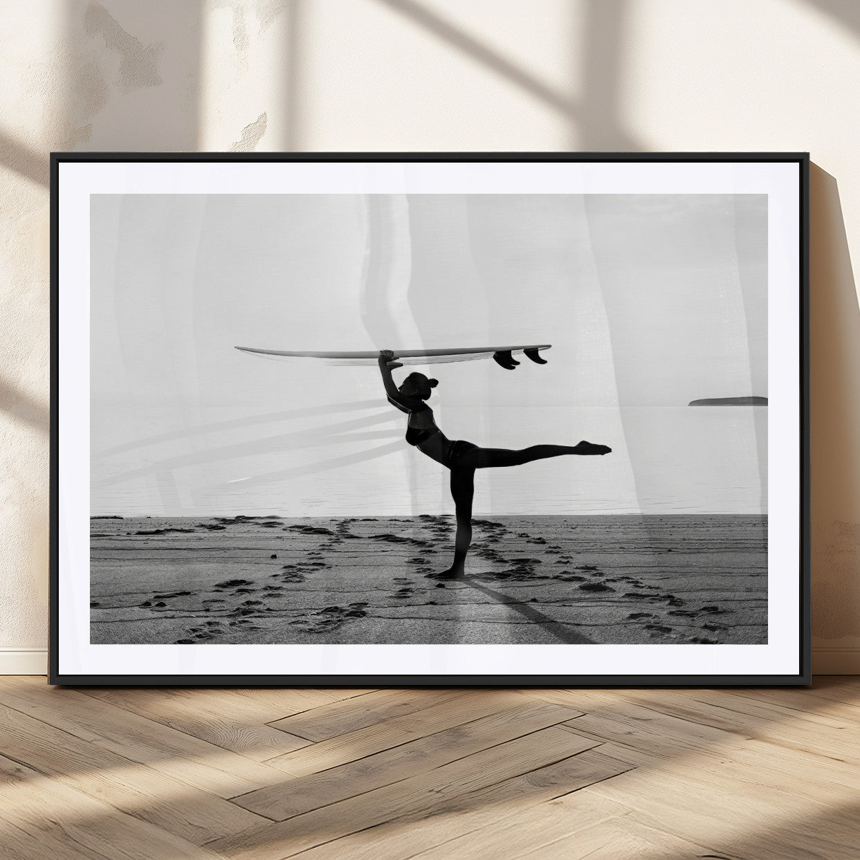A woman balances a surfboard on her head while striking a yoga pose on the beach, silhouetted against a calm ocean horizon.