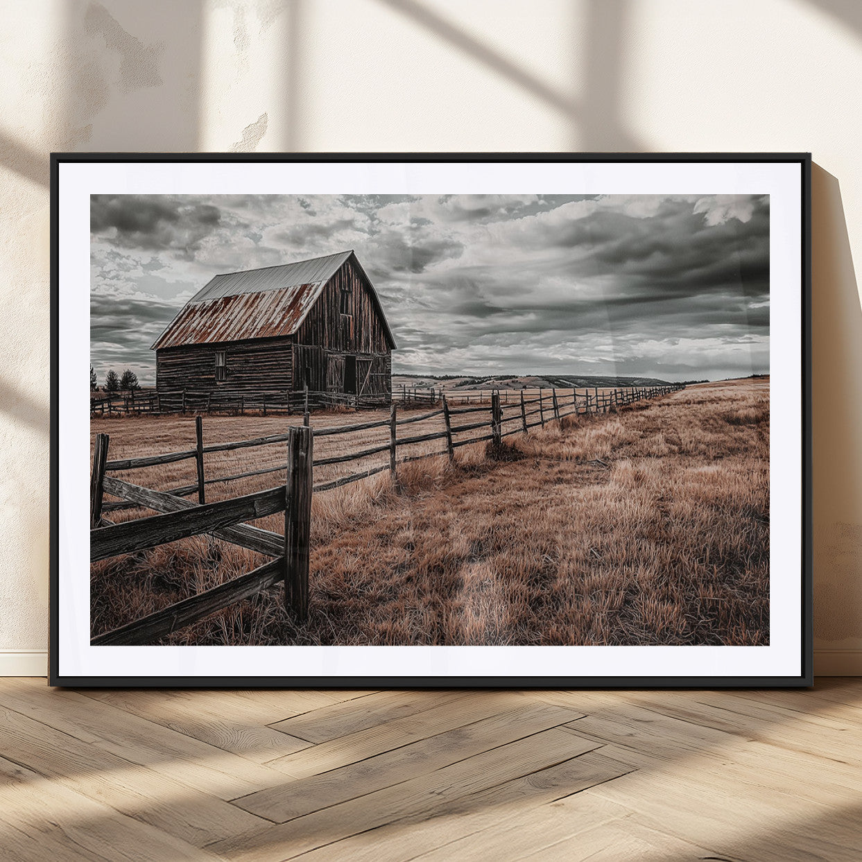 "Black-and-white photograph of a rustic barn with a weathered roof, surrounded by a wooden fence and vast golden fields under a moody sky."