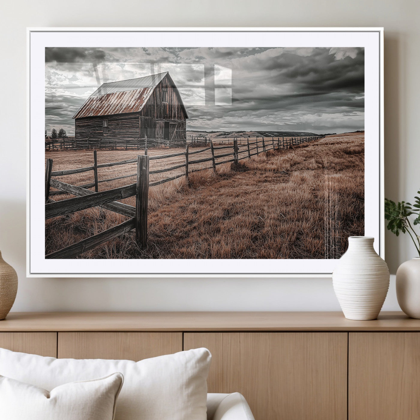 "Black-and-white photograph of a rustic barn with a weathered roof, surrounded by a wooden fence and vast golden fields under a moody sky."