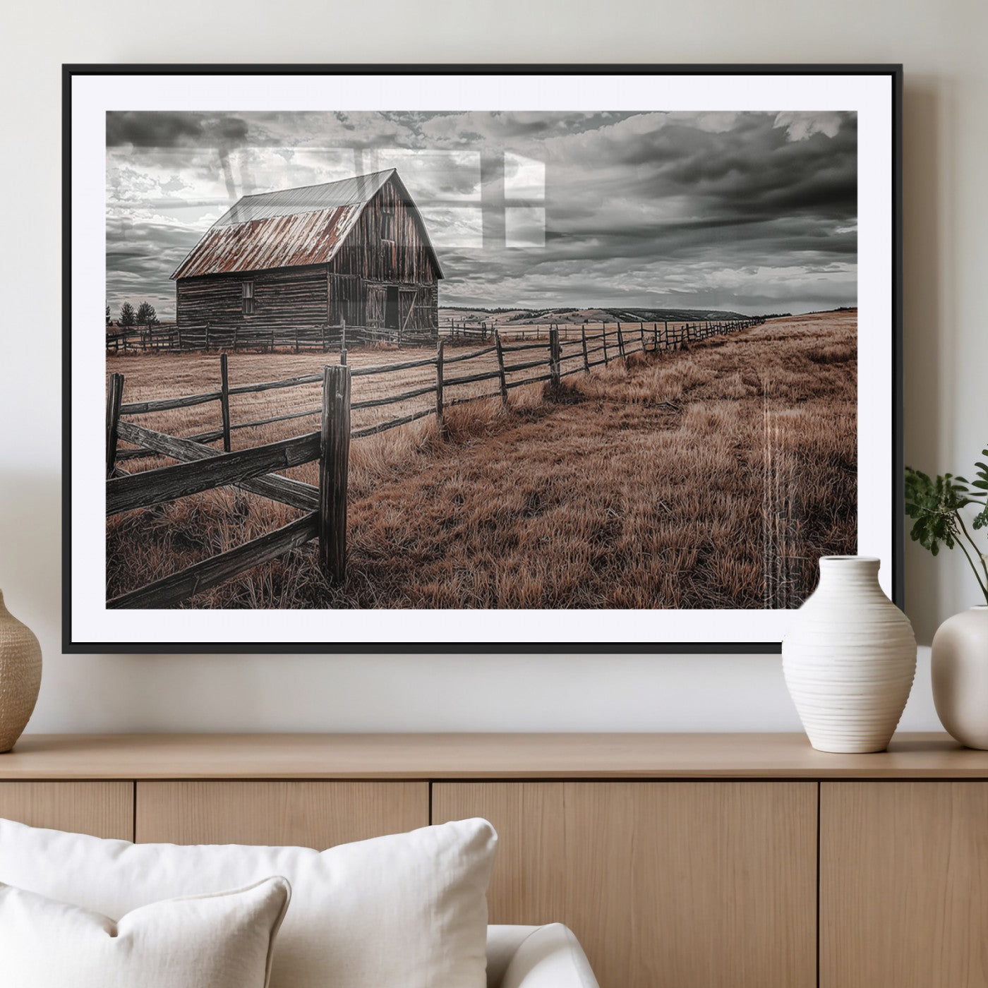 "Black-and-white photograph of a rustic barn with a weathered roof, surrounded by a wooden fence and vast golden fields under a moody sky."