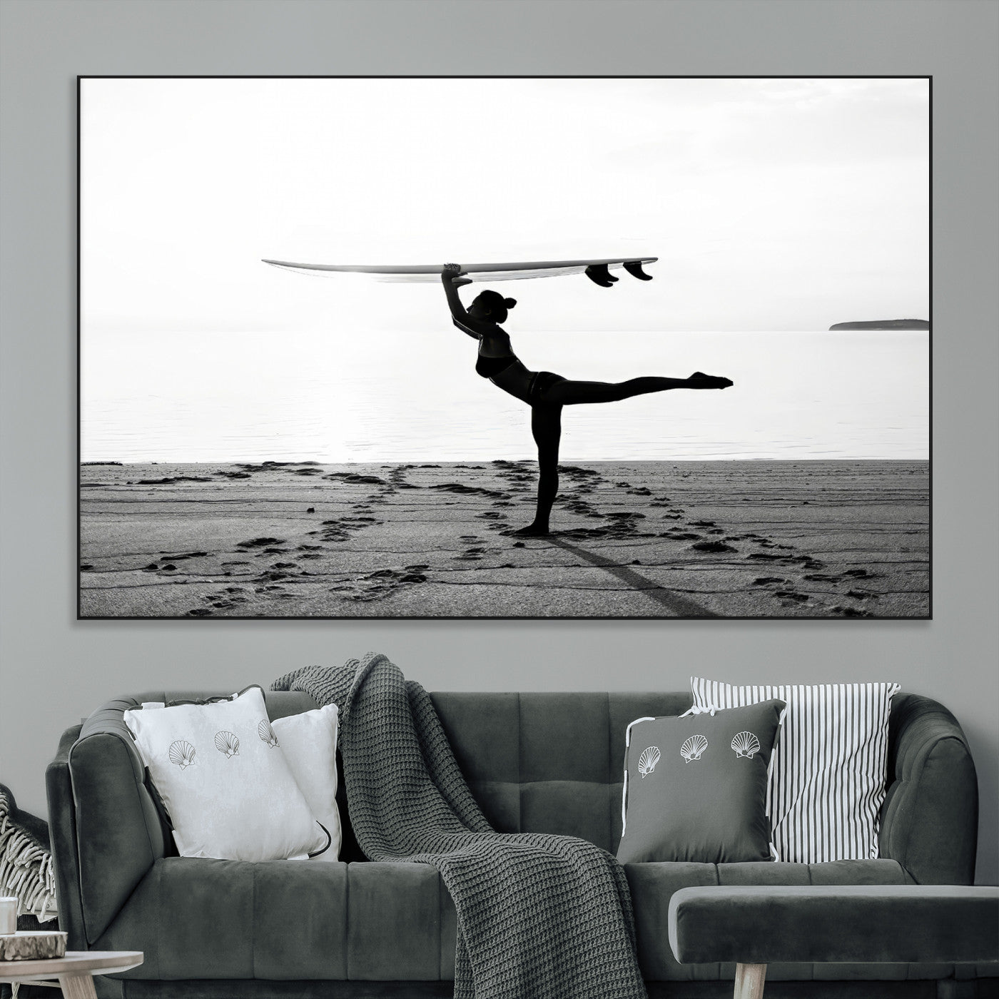 A woman balances a surfboard on her head while striking a yoga pose on the beach, silhouetted against a calm ocean horizon.