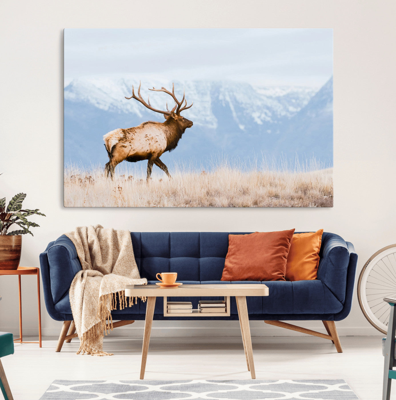 An elk walking on snowy grassland with majestic mountains in the background, ideal for rustic lodge or cabin wall decor.