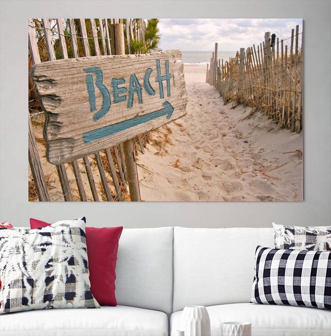 The Beach Is Calling You Wall Art Canvas Print features a sandy beach path with a wooden beach sign and arrow pointing to the ocean, beautifully displayed on museum-quality canvases.