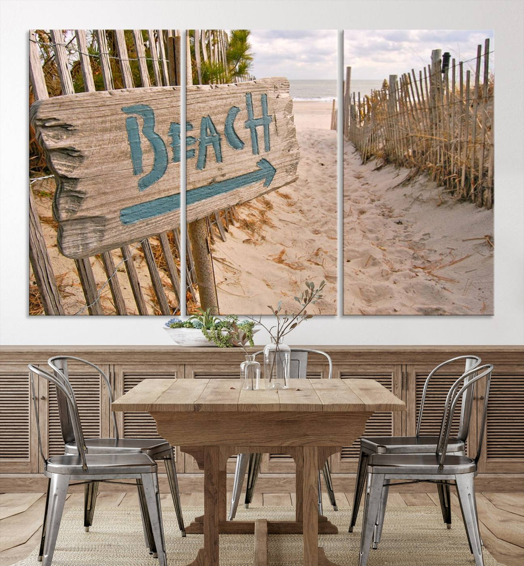 The Beach Is Calling You Wall Art Canvas Print features a sandy beach path with a wooden beach sign and arrow pointing to the ocean, beautifully displayed on museum-quality canvases.