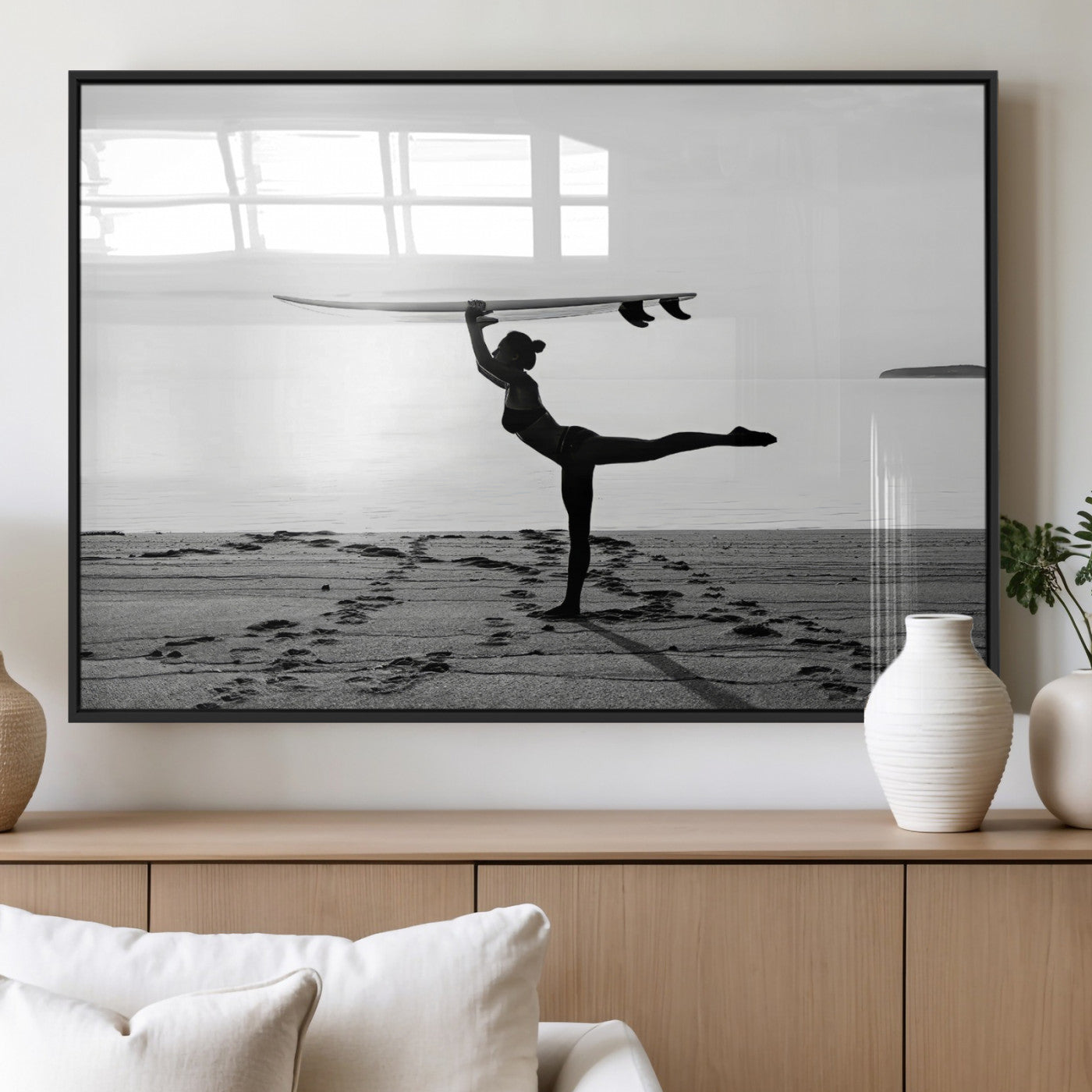 A woman balances a surfboard on her head while striking a yoga pose on the beach, silhouetted against a calm ocean horizon.