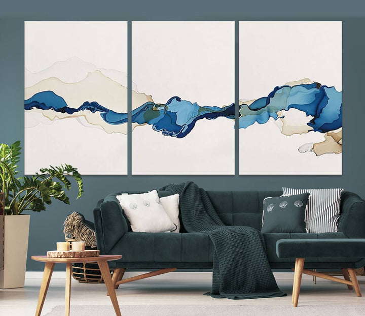 The modern living room showcases a set of three canvas prints with abstract blue art on museum-quality materials.