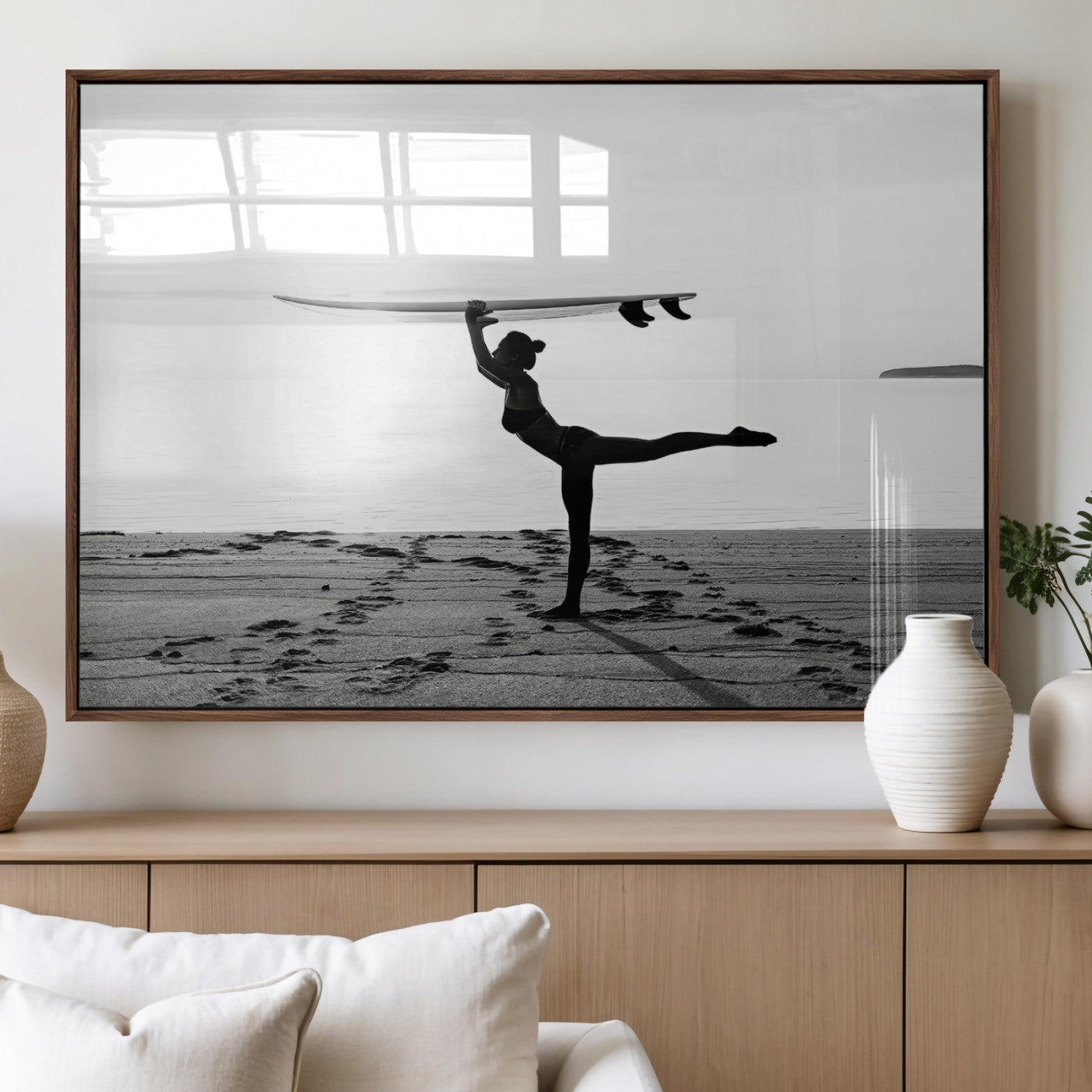 A woman balances a surfboard on her head while striking a yoga pose on the beach, silhouetted against a calm ocean horizon.