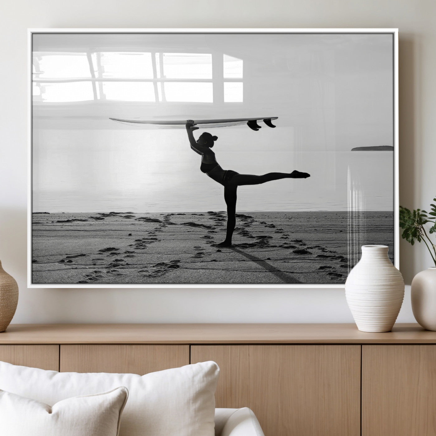 A woman balances a surfboard on her head while striking a yoga pose on the beach, silhouetted against a calm ocean horizon.