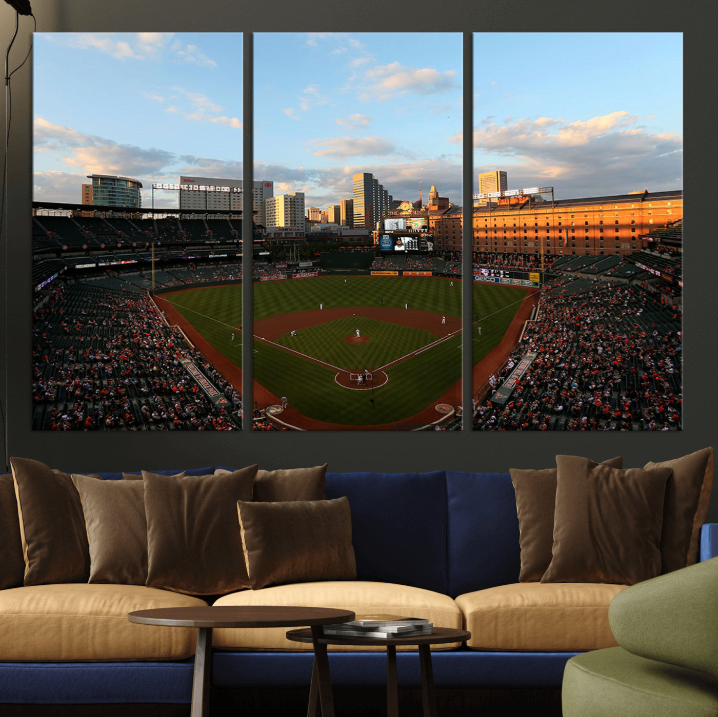 Aerial view of a baseball stadium at sunset with players on the field, fans in the stands, a city skyline in the background, and the setting sun casting warm light on the buildings.