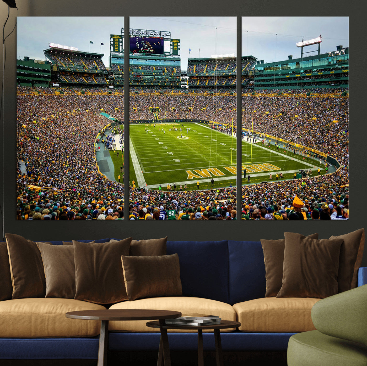Packed football stadium with thousands of fans watching an American football game on a well-maintained field. The scoreboard displays "Lambeau Field," and players are lined up on the field. The stadium is surrounded by multiple tiers of seating filled wit