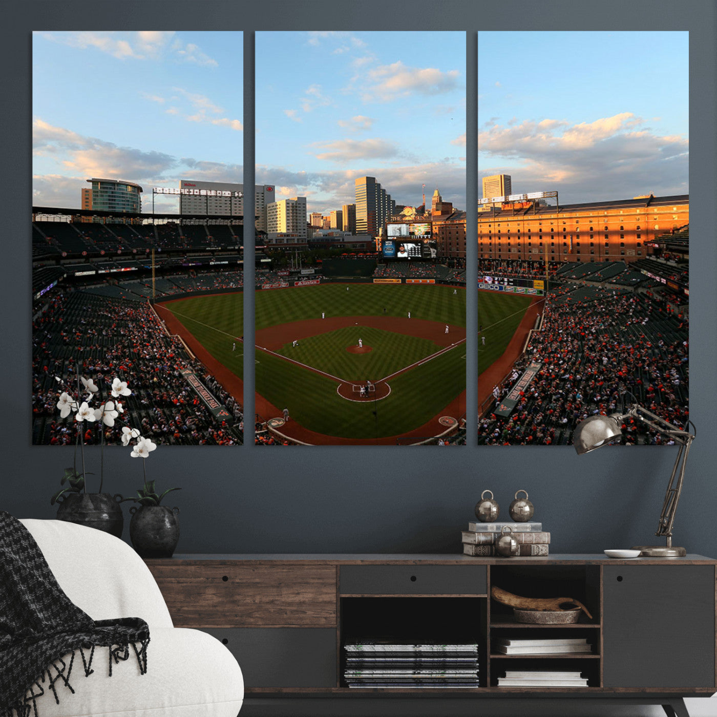 Aerial view of a baseball stadium at sunset with players on the field, fans in the stands, a city skyline in the background, and the setting sun casting warm light on the buildings.