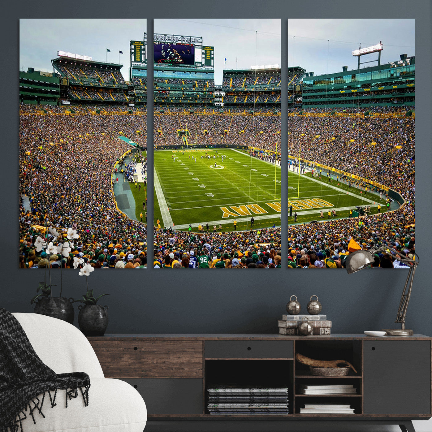 Packed football stadium with thousands of fans watching an American football game on a well-maintained field. The scoreboard displays "Lambeau Field," and players are lined up on the field. The stadium is surrounded by multiple tiers of seating filled wit