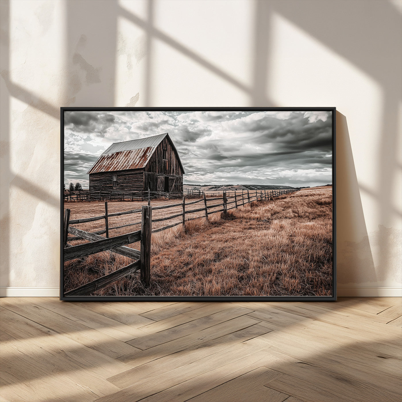 "Black-and-white photograph of a rustic barn with a weathered roof, surrounded by a wooden fence and vast golden fields under a moody sky."