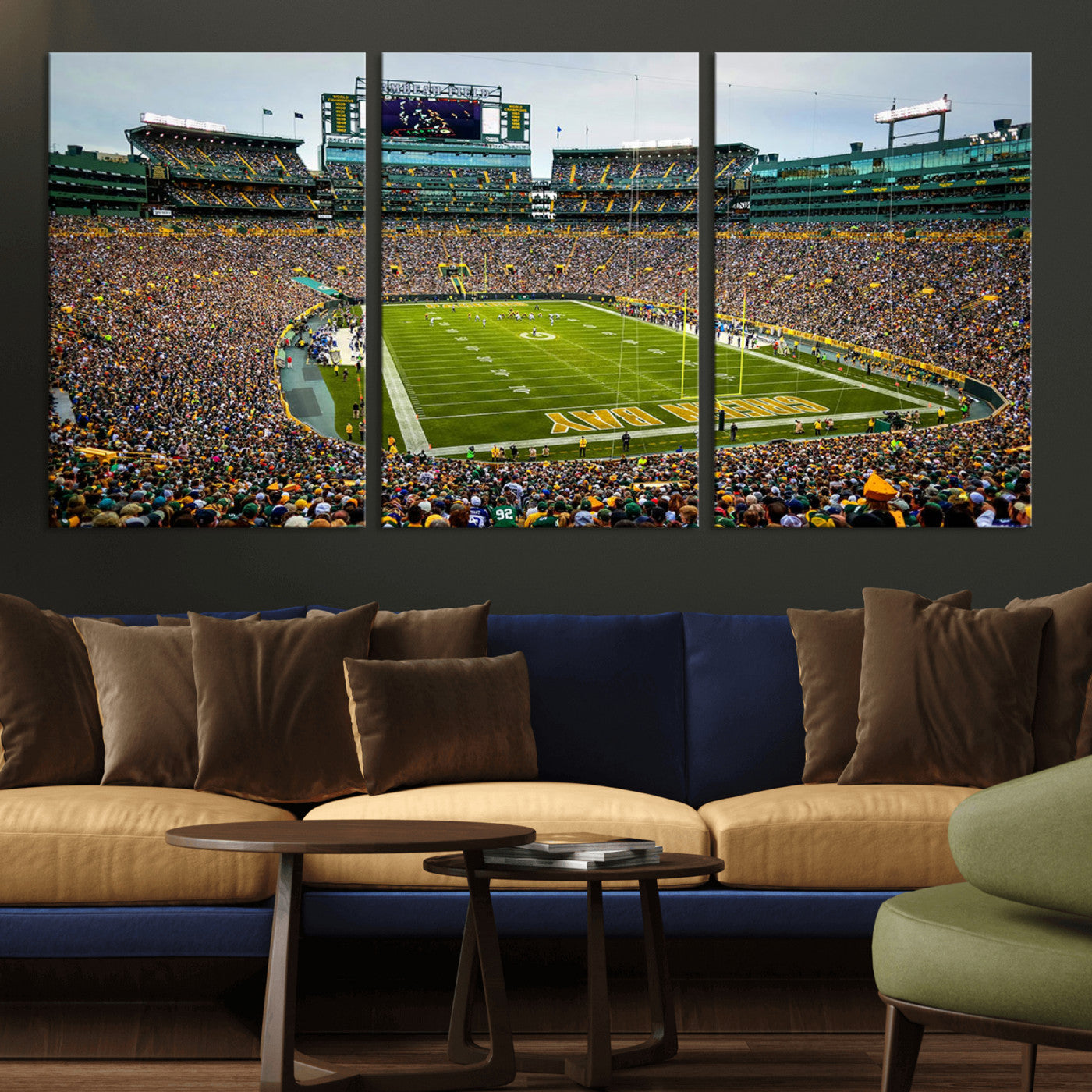 Packed football stadium with thousands of fans watching an American football game on a well-maintained field. The scoreboard displays "Lambeau Field," and players are lined up on the field. The stadium is surrounded by multiple tiers of seating filled wit