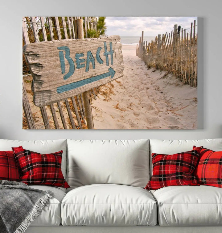 The Beach Is Calling You Wall Art Canvas Print features a sandy beach path with a wooden beach sign and arrow pointing to the ocean, beautifully displayed on museum-quality canvases.