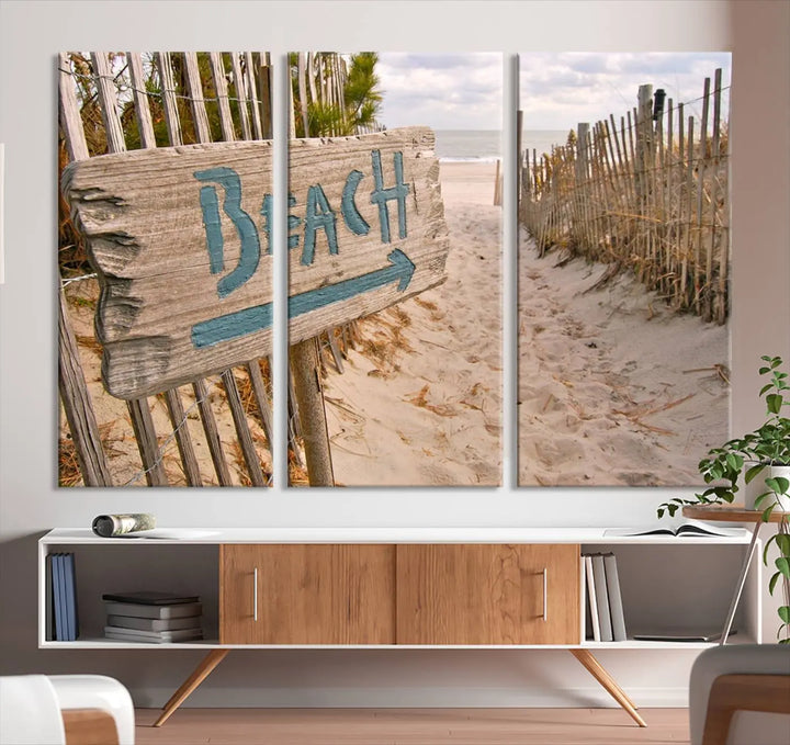 The Beach Is Calling You Wall Art Canvas Print features a sandy beach path with a wooden beach sign and arrow pointing to the ocean, beautifully displayed on museum-quality canvases.