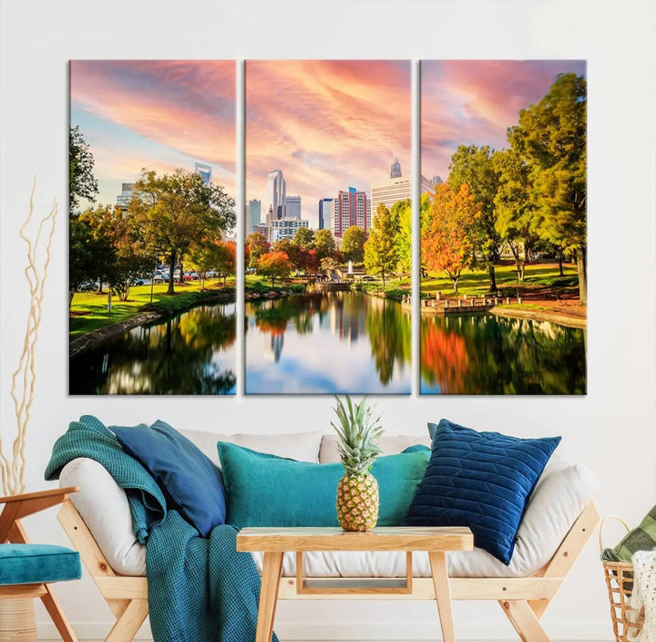 The "Charlotte City Park Sunset Pink and Orange Skyline Canvas Print" is elegantly displayed in a modern living room. Crafted on museum-quality canvas, this triptych wall art showcases a cityscape and park at sunset, complete with a UV-protective coating, and is ready to hang.