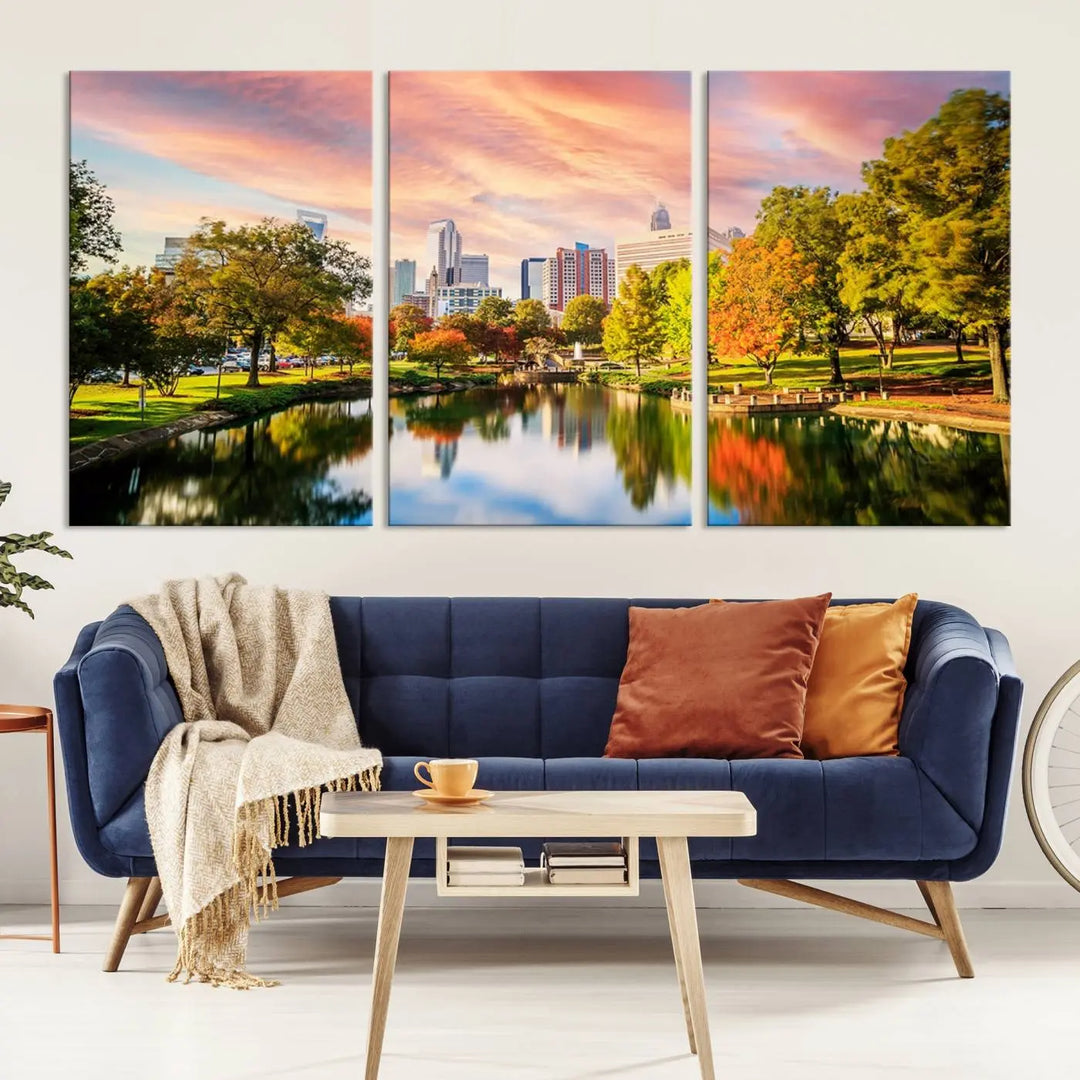 The "Charlotte City Park Sunset Pink and Orange Skyline Canvas Print" is elegantly displayed in a modern living room. Crafted on museum-quality canvas, this triptych wall art showcases a cityscape and park at sunset, complete with a UV-protective coating, and is ready to hang.