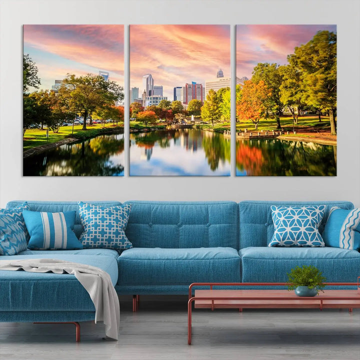 The "Charlotte City Park Sunset Pink and Orange Skyline Canvas Print" is elegantly displayed in a modern living room. Crafted on museum-quality canvas, this triptych wall art showcases a cityscape and park at sunset, complete with a UV-protective coating, and is ready to hang.