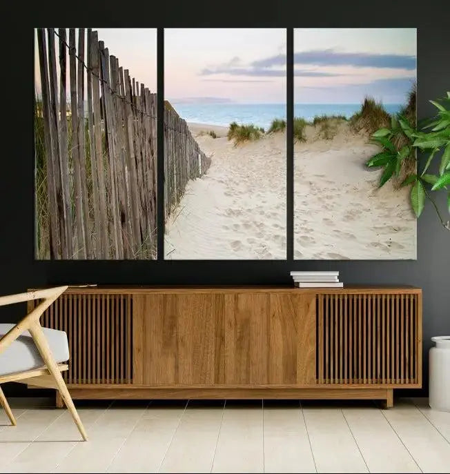 The Coastal Beach Fence Wall Art Ocean Landscape Canvas Print, featuring a triptych of a sandy beach scene with dunes and fences, is gallery wrapped on museum-quality canvas and displayed on a modern dark wall.