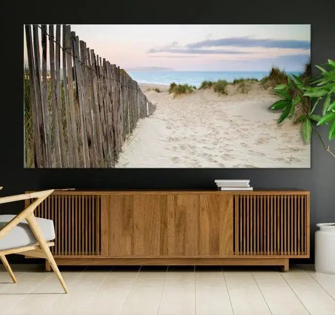 The Coastal Beach Fence Wall Art Ocean Landscape Canvas Print, featuring a triptych of a sandy beach scene with dunes and fences, is gallery wrapped on museum-quality canvas and displayed on a modern dark wall.