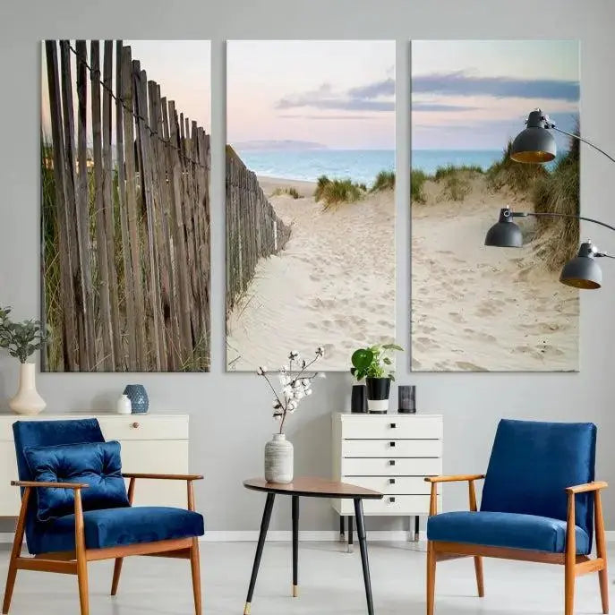 The Coastal Beach Fence Wall Art Ocean Landscape Canvas Print, featuring a triptych of a sandy beach scene with dunes and fences, is gallery wrapped on museum-quality canvas and displayed on a modern dark wall.