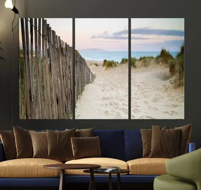 The Coastal Beach Fence Wall Art Ocean Landscape Canvas Print, featuring a triptych of a sandy beach scene with dunes and fences, is gallery wrapped on museum-quality canvas and displayed on a modern dark wall.