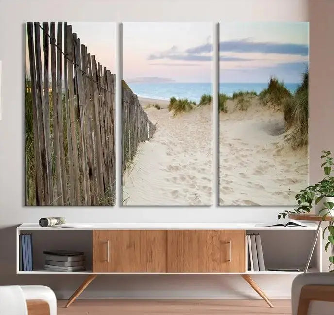 The Coastal Beach Fence Wall Art Ocean Landscape Canvas Print, featuring a triptych of a sandy beach scene with dunes and fences, is gallery wrapped on museum-quality canvas and displayed on a modern dark wall.