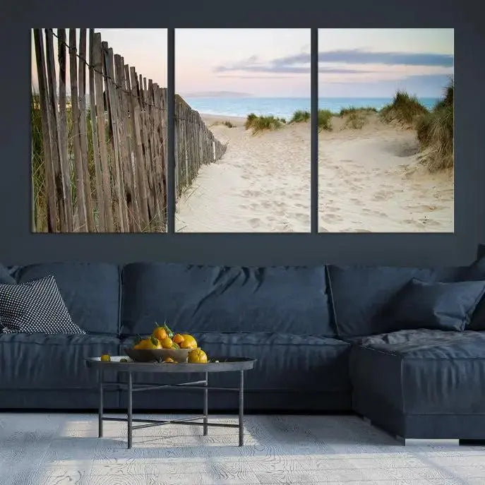 The Coastal Beach Fence Wall Art Ocean Landscape Canvas Print, featuring a triptych of a sandy beach scene with dunes and fences, is gallery wrapped on museum-quality canvas and displayed on a modern dark wall.