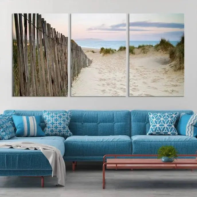 The Coastal Beach Fence Wall Art Ocean Landscape Canvas Print, featuring a triptych of a sandy beach scene with dunes and fences, is gallery wrapped on museum-quality canvas and displayed on a modern dark wall.