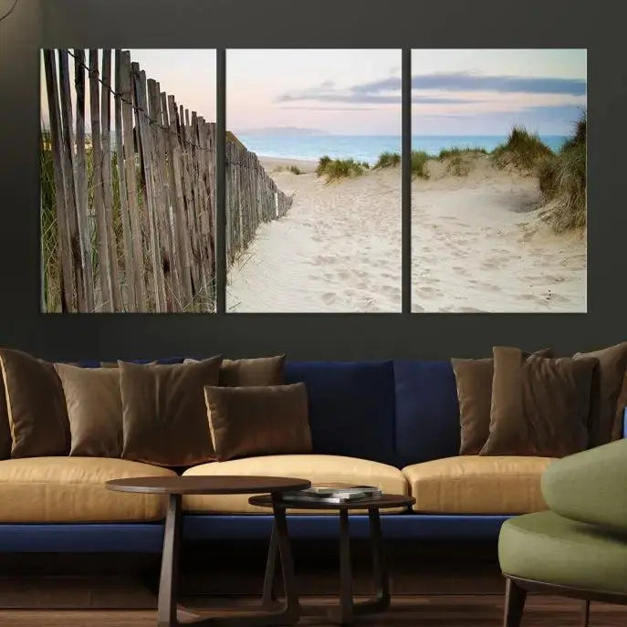 The Coastal Beach Fence Wall Art Ocean Landscape Canvas Print, featuring a triptych of a sandy beach scene with dunes and fences, is gallery wrapped on museum-quality canvas and displayed on a modern dark wall.