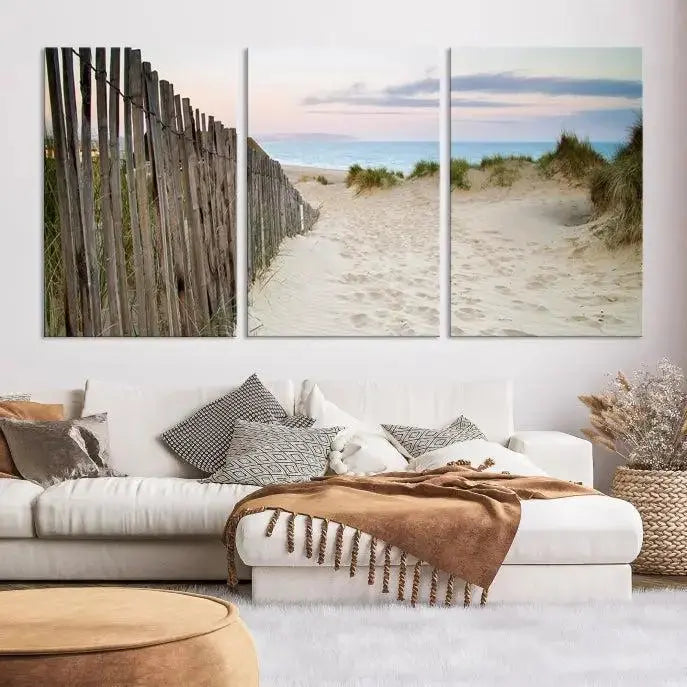 The Coastal Beach Fence Wall Art Ocean Landscape Canvas Print, featuring a triptych of a sandy beach scene with dunes and fences, is gallery wrapped on museum-quality canvas and displayed on a modern dark wall.