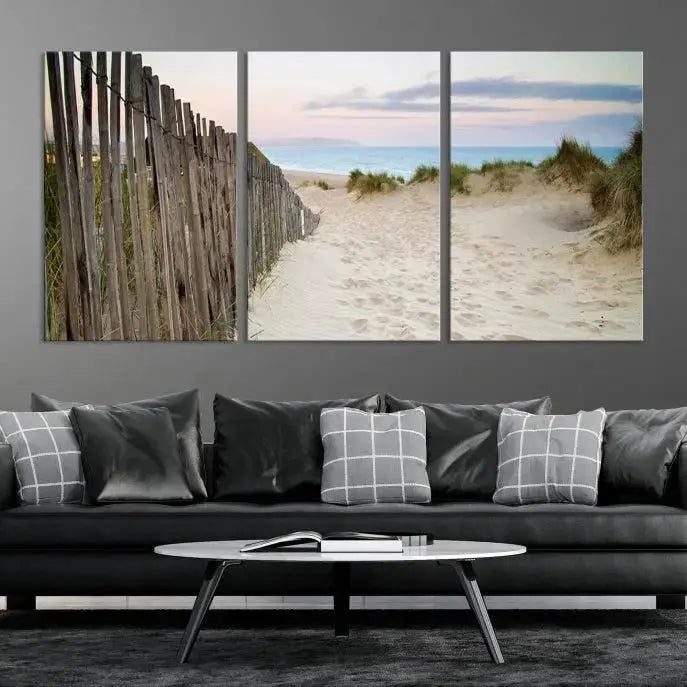 The Coastal Beach Fence Wall Art Ocean Landscape Canvas Print, featuring a triptych of a sandy beach scene with dunes and fences, is gallery wrapped on museum-quality canvas and displayed on a modern dark wall.