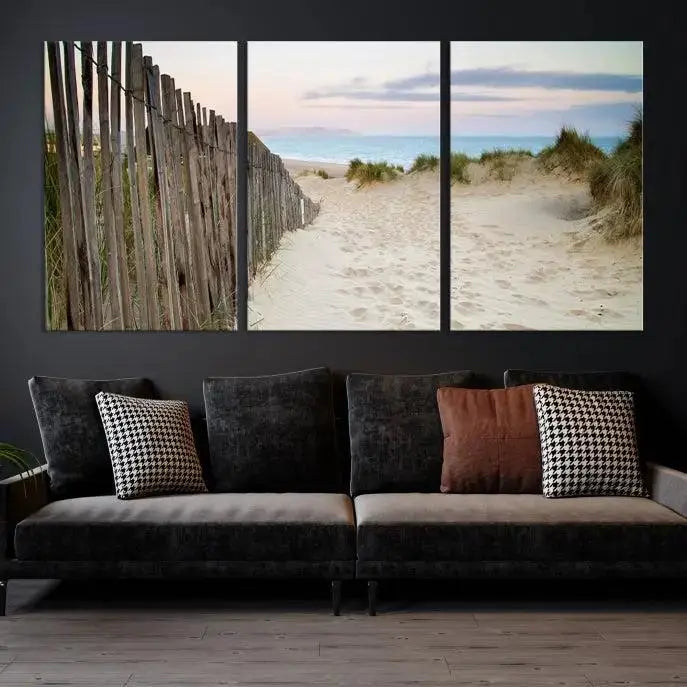 The Coastal Beach Fence Wall Art Ocean Landscape Canvas Print, featuring a triptych of a sandy beach scene with dunes and fences, is gallery wrapped on museum-quality canvas and displayed on a modern dark wall.