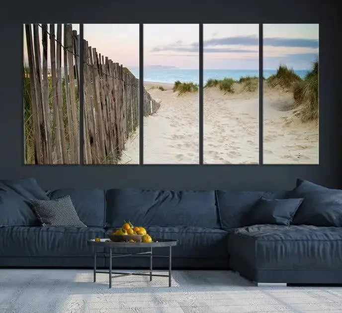 The Coastal Beach Fence Wall Art Ocean Landscape Canvas Print, featuring a triptych of a sandy beach scene with dunes and fences, is gallery wrapped on museum-quality canvas and displayed on a modern dark wall.