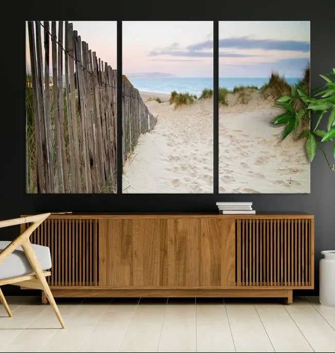 The Coastal Beach Fence Wall Art Ocean Landscape Canvas Print, featuring a triptych of a sandy beach scene with dunes and fences, is gallery wrapped on museum-quality canvas and displayed on a modern dark wall.