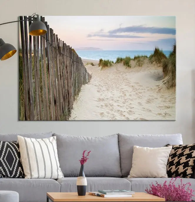 The Coastal Beach Fence Wall Art Ocean Landscape Canvas Print, featuring a triptych of a sandy beach scene with dunes and fences, is gallery wrapped on museum-quality canvas and displayed on a modern dark wall.