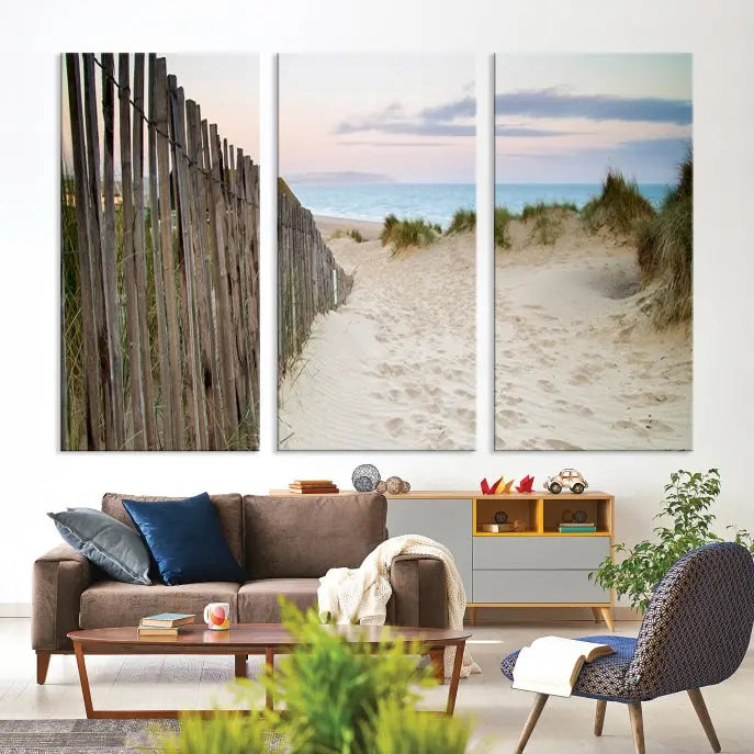 The Coastal Beach Fence Wall Art Ocean Landscape Canvas Print, featuring a triptych of a sandy beach scene with dunes and fences, is gallery wrapped on museum-quality canvas and displayed on a modern dark wall.