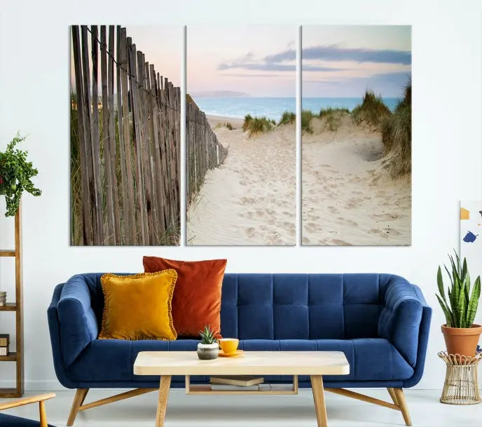 The Coastal Beach Fence Wall Art Ocean Landscape Canvas Print, featuring a triptych of a sandy beach scene with dunes and fences, is gallery wrapped on museum-quality canvas and displayed on a modern dark wall.