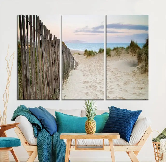 The Coastal Beach Fence Wall Art Ocean Landscape Canvas Print, featuring a triptych of a sandy beach scene with dunes and fences, is gallery wrapped on museum-quality canvas and displayed on a modern dark wall.