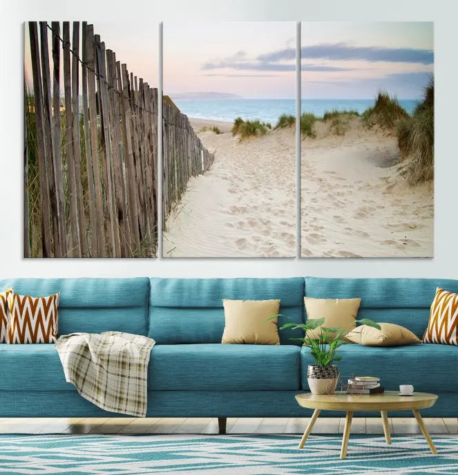 The Coastal Beach Fence Wall Art Ocean Landscape Canvas Print, featuring a triptych of a sandy beach scene with dunes and fences, is gallery wrapped on museum-quality canvas and displayed on a modern dark wall.